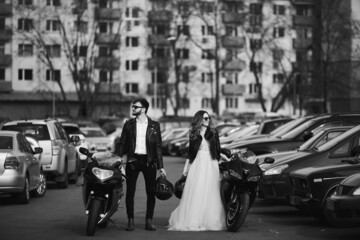 Stylish bride and bearded groom stand beside sports motorcycles on the urban street. A beautiful couple of newlyweds posing with their motorbikes - Powered by Adobe