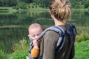 Caucasian mother with a baby carrier taking a walk around a lake in the middle of nature. healthy...