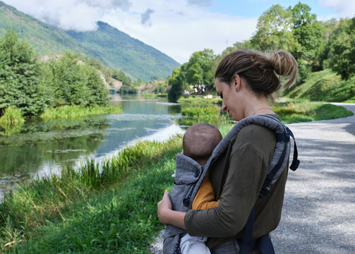 Caucasian Mother With A Baby Carrier Taking A Walk Around A Lake In The Middle Of Nature. Healthy Lifestyle