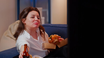 Woman spending free time with takeaway food and TV while sitting in living room. Caucasian adult eating hamburger and drinking beer, looking at television after work. Person enjoying meal