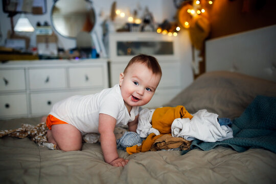 funny european chubby baby crawling among clothes on bed, children's clothes and conscious consumption, lot of clothes and child, big pile of clean clothes and baby playing, childhood and emotions