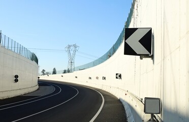 Road curve at the exit of an underpass with high white concrete wall and directional arrow signs....