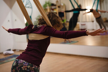 Młoda kobieta z kręconymi włosami prowadzi zajęcia z jogi na szarfach, w lustrze widać odbicie innych uczestniczek zajęć