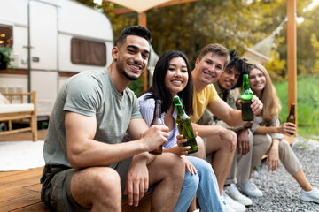 Diverse young friends having party near RV outdoors, drinking beer, communicating and laughing on autumn camping trip