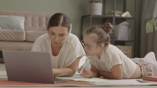Slowmo shot of mom and daughter studying on laptop together lying casually on floor in living room