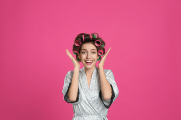 Emotional young woman in silk bathrobe with hair curlers on pink background