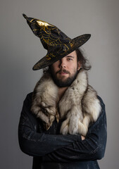 A young dark-haired man in a witch's hat, with his arms folded on his chest.