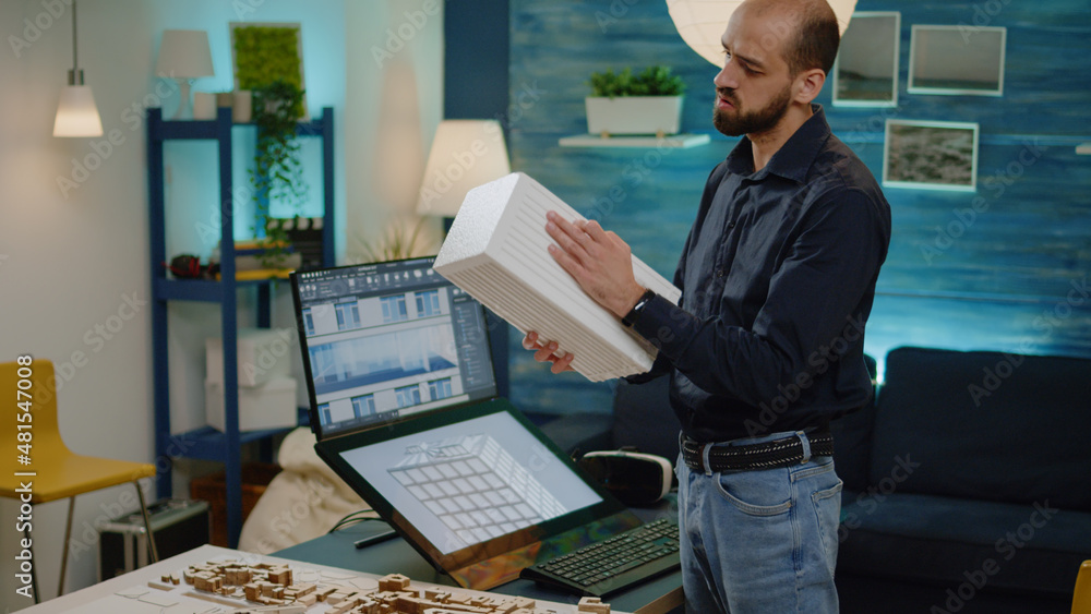 Poster Man working as architect analyzing maquette in hand. Young engineer checking building model with blueprint plan on computer monitor for construction layout and development project