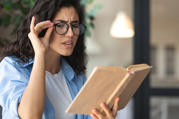 Beautiful young woman in glasses reading book, having poor eyesight at home