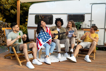 Group of cool diverse friends with American flag, beer and guitar having student party near RV on...