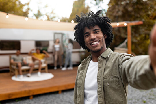Funky black guy taking selfie near RV, resting with diverse friends outdoors, enjoying autumn camping trip, copy space