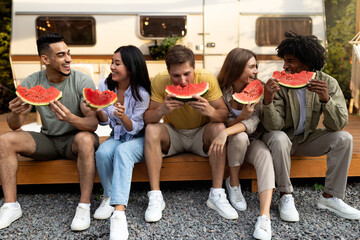Cheerful diverse young friends eating fresh juicy watermelon, sitting near camper van on autumn...