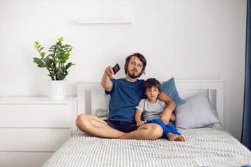 bearded father and son are sitting on the bed in T-shirts and watching football match on TV. a man holds a remote control in his hands to switch channels