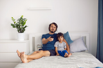 bearded father and son are sitting on the bed in T-shirts and watching football match on TV. a man holds a remote control in his hands to switch channels