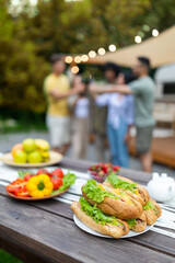 Sandwiches and vegetables, picnic foods on table outdoors, group of young friends toasting with beer on background