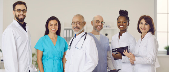 Happy clinic or hospital staff at work. Multiracial team of friendly doctors, including clinicians and cardiologists, in scrubs and white coat uniforms standing together, looking at camera and smiling
