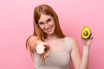Young redhead woman holding an avocado isolated on pink background pointing front with happy expression