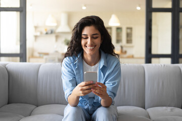 Obraz premium Happy young woman sitting on couch with smartphone, checking social media, browsing internet, chatting online from home