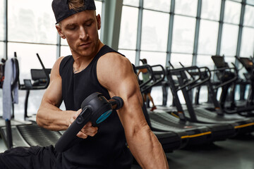 Obraz premium A young man is using and enjoying percussion massage gun to relax and relief the pain in body and muscles in the gym