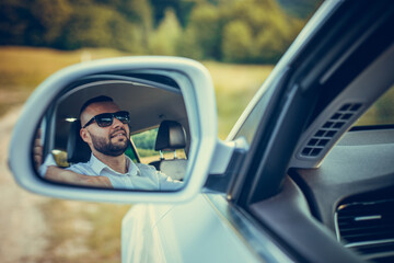 Bearded caucasian business man looking at camera through rear view mirror. Head and shoulders. Summer background.