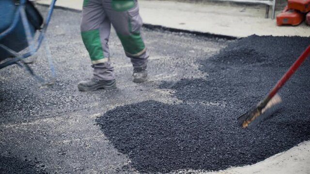 Asphalt laborers level fresh asphalt on a road construction site using an asphalt lute, shovel, and a wheelbarrow.