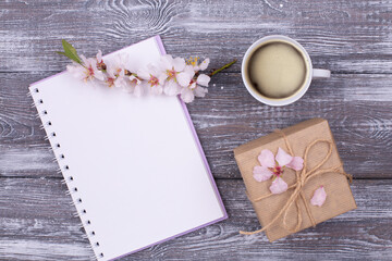 Spring composition with empty diary, cup of coffee, gift wrapped in kraft paper, spring blooming almond branch with flowers on a gray wooden table. Flat lay, copy space
