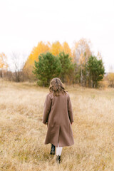 Beautiful stylish woman with windy hair in wool coat posing among wild grass. Carefree moment, stylish image, walking on autumn nature. Fashionable young female standing on windy coast.