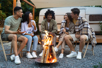 Multiracial friends sitting near bonfire, talking to each other, laughing, spending autumn evening...