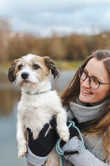 Junge lächenlde Frau hält einen kleinen Terrier Hund auf ihren Armen an einem See. Freundschaft, Zusammenhalt.