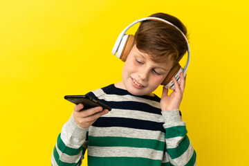 Little redhead boy isolated on yellow background listening music with a mobile and singing