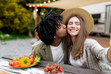 Loving Afro guy kissing his Caucasian girlfriend while making selfie on camping trip in autumn