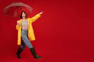 Excited woman in yellow waterproof raincoat and boots walking with umbrella and pointing at free space on red background