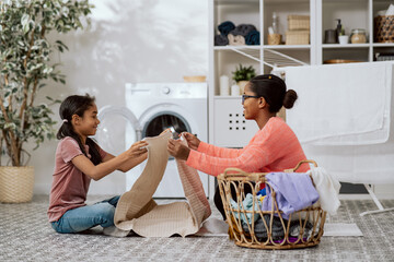Mother and daughter are sitting on floor in home laundry room, the girl is helping woman with household chores, they are putting together clean washed clothes taken out of a large wicker basket