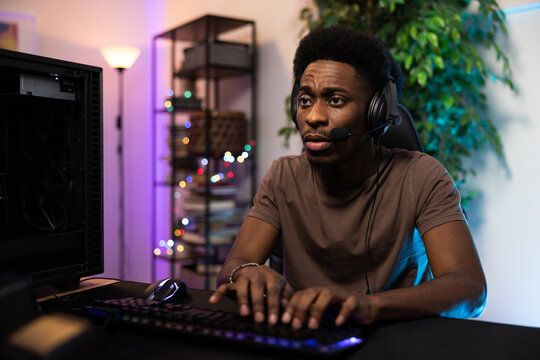 Portrait of professional video gamer afro-American man is focused on winning round he has headset backlit keyboard and mouse to play the computer, purple and blue neon lights in the room