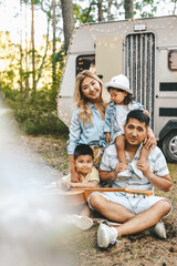 An Asian multiracial family with two children are camping in nature on a road trip. Dad plays guitar to mom daughter and son in the woods in the summer. Selective focus