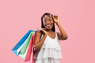 Excited black lady holding shopping bags, touching sunglasses, buying goods on big sale over pink background