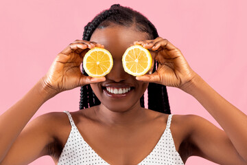 Vitamins for beauty and skin care. Positive Afro woman covering both eyes with lemon halves on pink background
