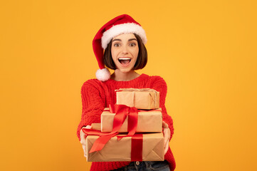 Emotional happy woman in santa hat holding gift boxes and stretching them to camera over yellow studio background