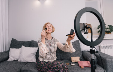 A photo of a beautiful elderly blogger woman sitting on the couch telling viewers online how to apply makeup while looking at the screen of a smartphone standing on a tripod.