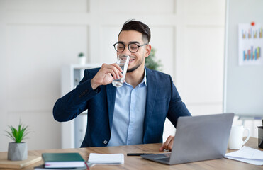 Cool Arab businessman in suit drinking water while working on laptop at modern office