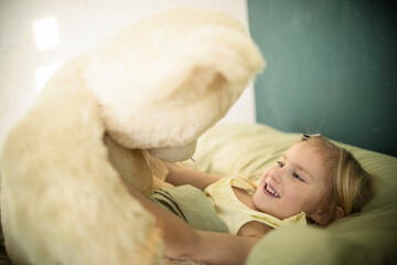 A little caucasian girl plays with a teddy bear in bed, hugs him and puts him to sleep