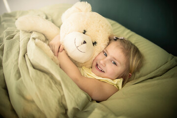 A little caucasian girl plays with a teddy bear in bed, hugs him and puts him to sleep