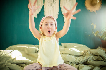 Little caucasian girl playing yoga and has fun on the bed in a bright sunny room
