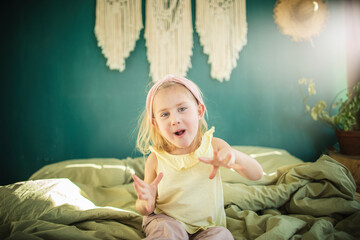 Little caucasian girl playing yoga and has fun on the bed in a bright sunny room