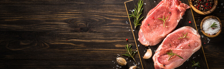 Fresh pork steaks with herbs and spices on craft board at wooden table Long banner format.