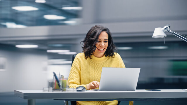 Modern Office: Happy Smiling Hispanic Businesswoman Sitting at Her Desk Working on a Laptop Computer Celebrates Victory. Latin Female Entrepreneur is a Happy Winner. Motion Blur Background