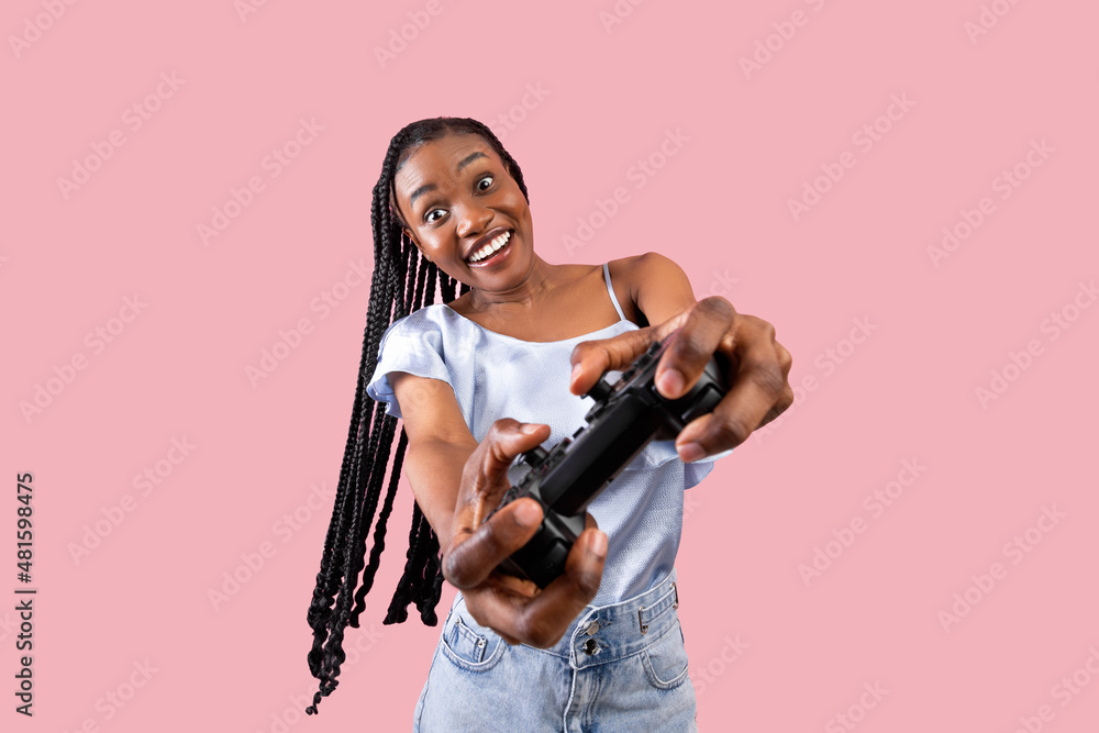 Wall mural Modern entertainments concept. Excited young black lady with joystick playing video game on pink studio background