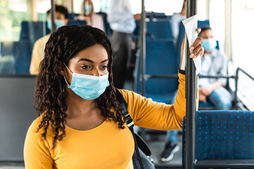 Black woman in surgical mask standing in bus using tissue