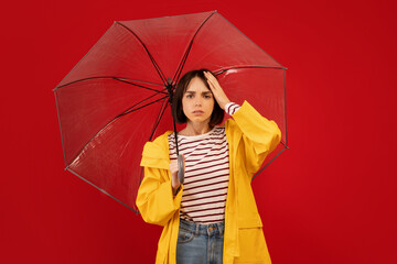 Weather dependence concept. Young woman touching head and standing under umbrella, posing over red background