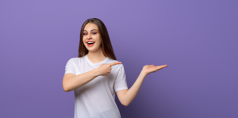 Portrait of young woman surprise gazing and pointing finger aside and show something in hand over purple background